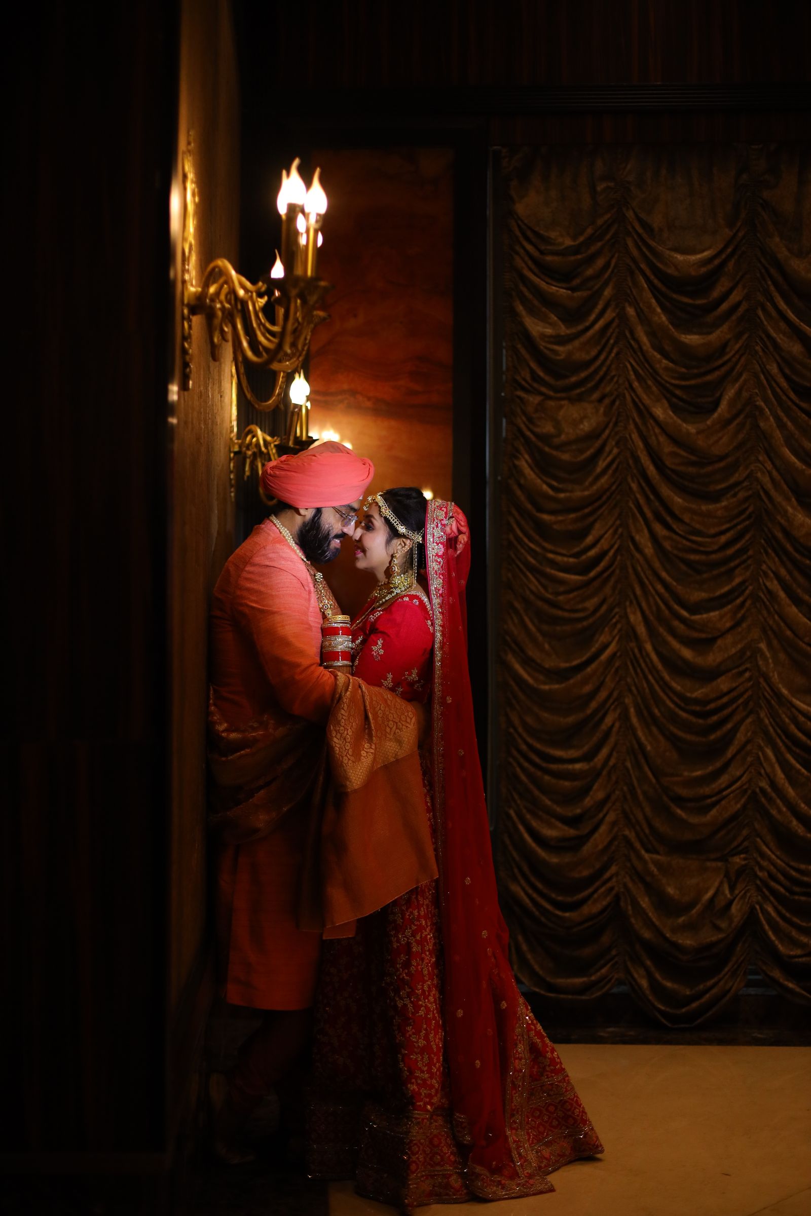 A couple stand forehead to forehead in a narrow doorway lit by five candles in a brass wall bracket, the panelled room around them nearly black.