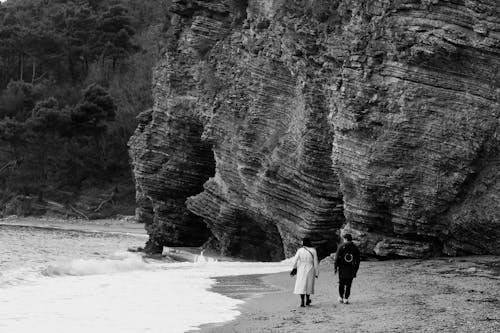 People Walking near Rocks on Sea Shore in Black and White
