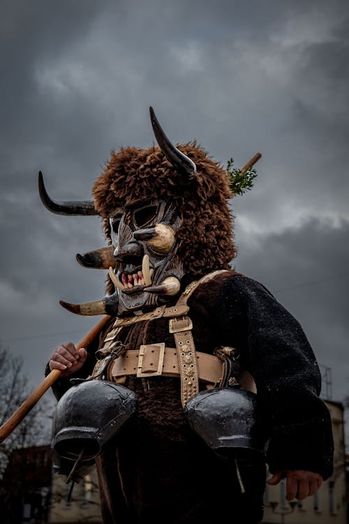 A Person Wearing a Costume with a Scary Mask with Horns Walking Outside under Dark Clouds 