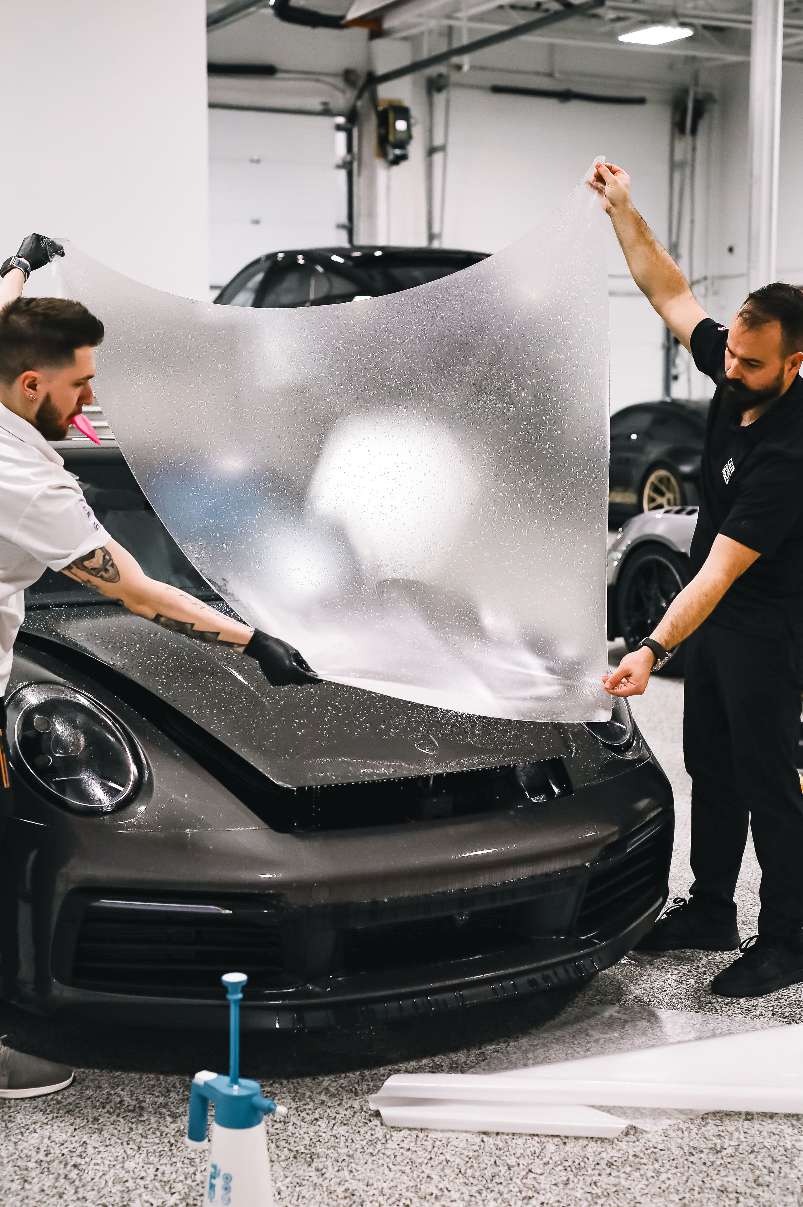 Two technicians applying a protective film on a sleek sports car in a garage setting.