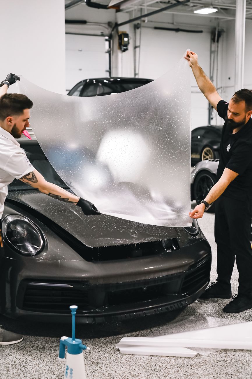 Two technicians applying a protective film on a sleek sports car in a garage setting.