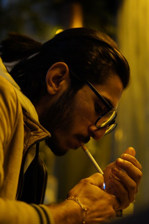 Free Profile shot of a man with glasses lighting a cigarette, capturing an intimate night moment. Stock Photo