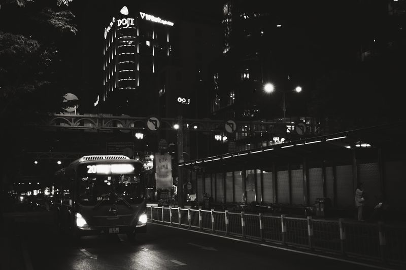 Black and white capture of a city street at night with a bus and illuminated buildings.
