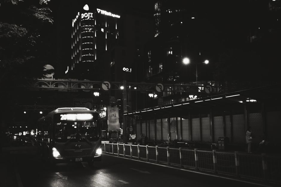 Black and white capture of a city street at night with a bus and illuminated buildings.