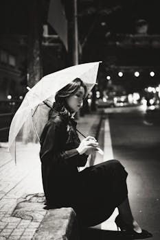 A stylish woman sits on a city curb holding an umbrella during a quiet urban night.