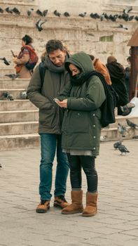 Couple in winter attire looking at smartphone on busy city street, surrounded by pigeons.