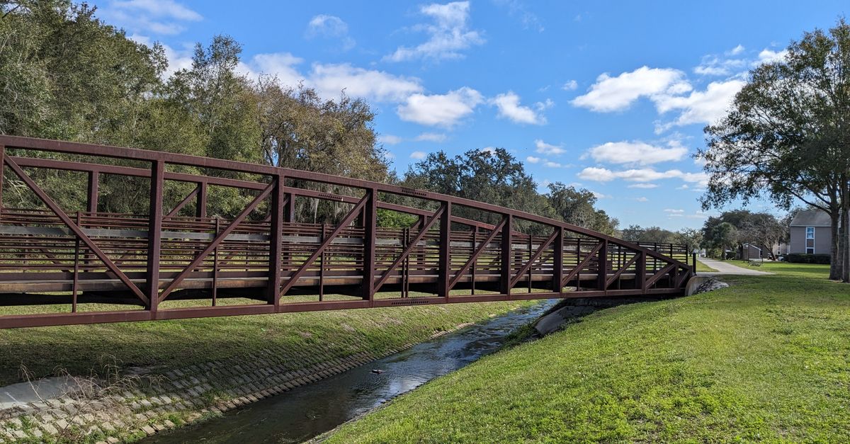 A rustic footbridge spans a quiet stream under a clear blue sky in Kissimmee, Florida.