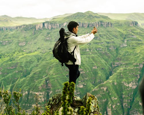 Man Standing on Rock Edge over Mountains and Taking Pictures