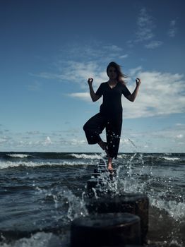 Silhouette of a woman balancing on a breakwater in the ocean with arms raised against a morning sky.