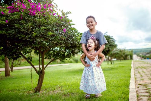 Free Smiling siblings in a park with vibrant greenery and colorful flowers, enjoying a sunny day outdoors. Stock Photo