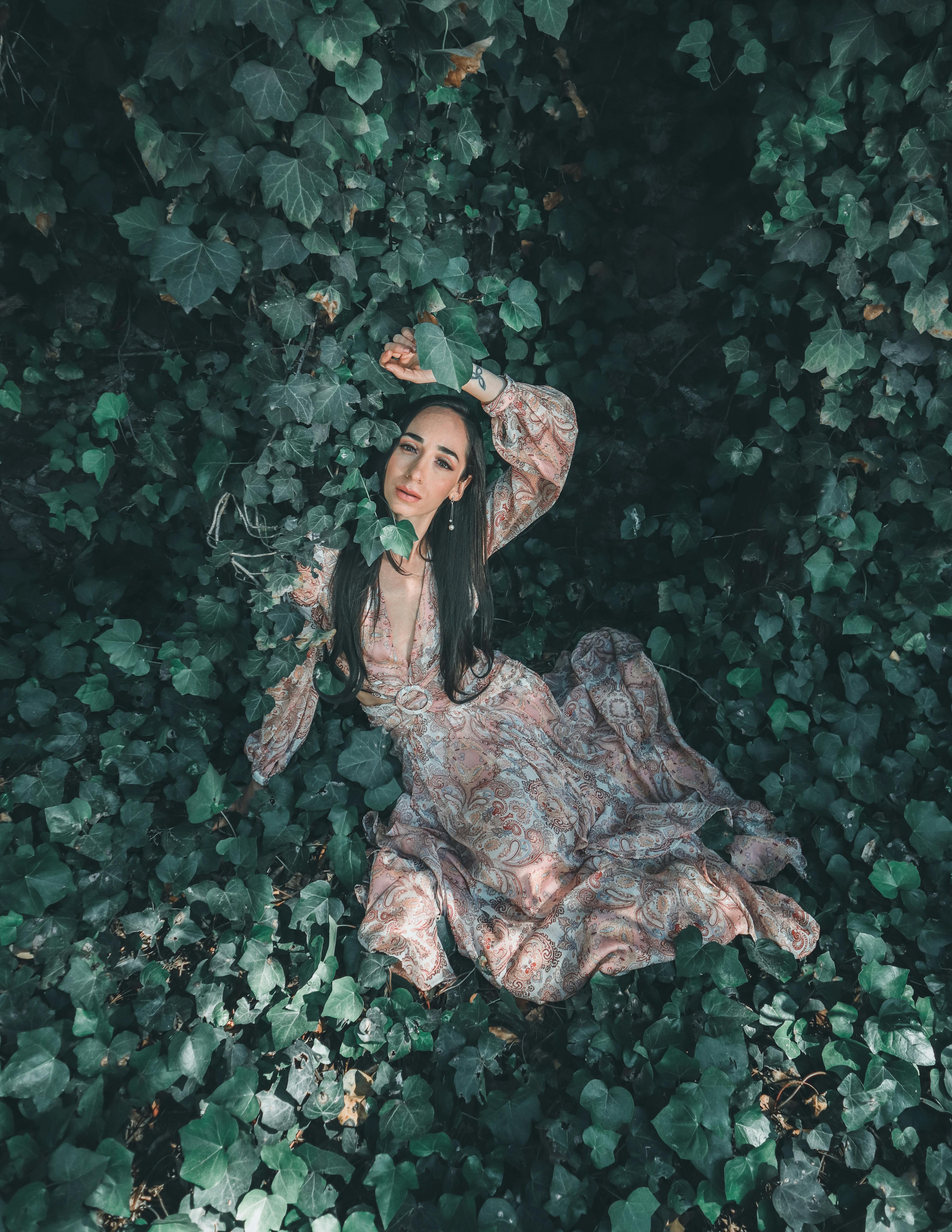 A woman in a floral dress lies on lush green leaves, creating a serene and artistic scene.