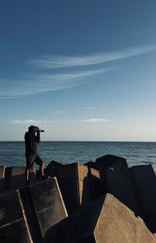 A photographer stands on concrete blocks, capturing the serene sea under a blue sky.