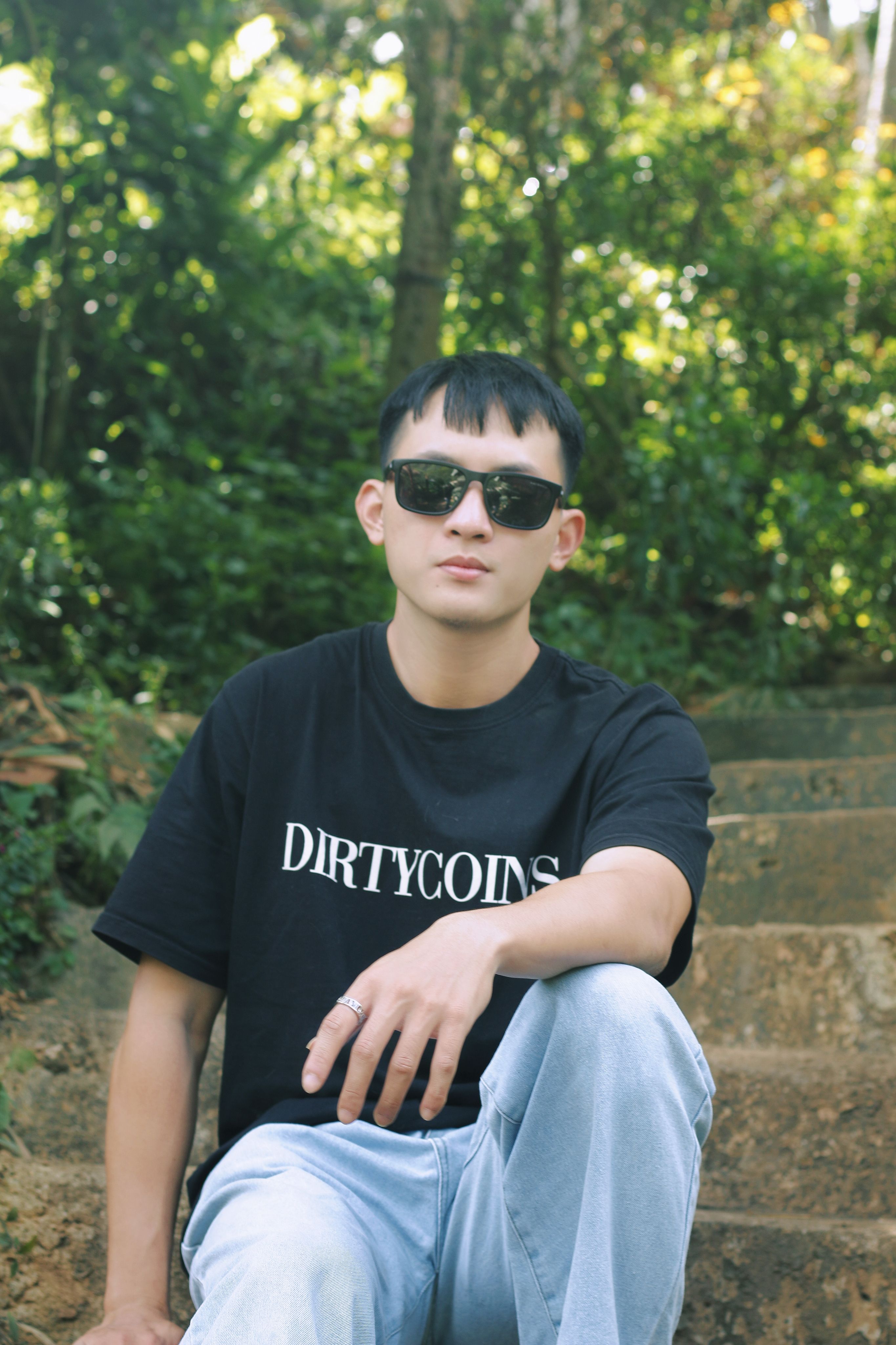 Casual young man in sunglasses and t-shirt relaxing on outdoor stone steps surrounded by greenery.