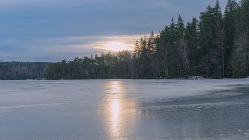 Free Serene winter scene featuring a frozen lake with the sunset reflecting on the icy surface surrounded by forest. Stock Photo