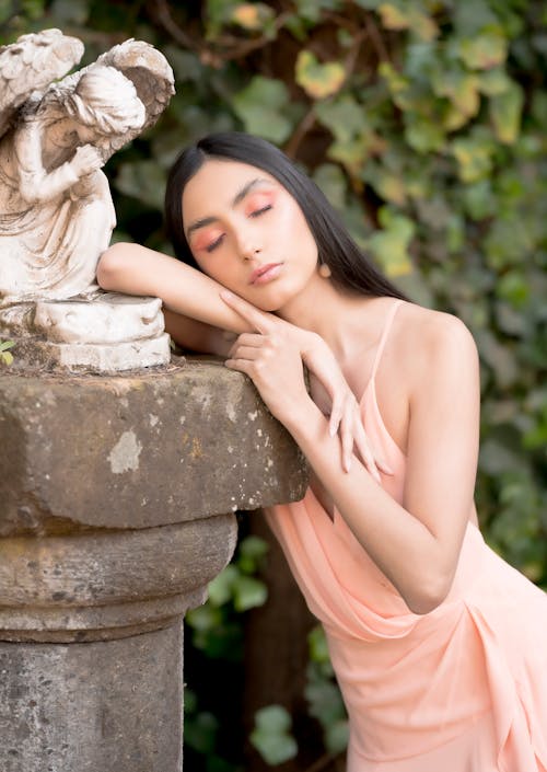 A woman in a peach dress is leaning against a stone statue