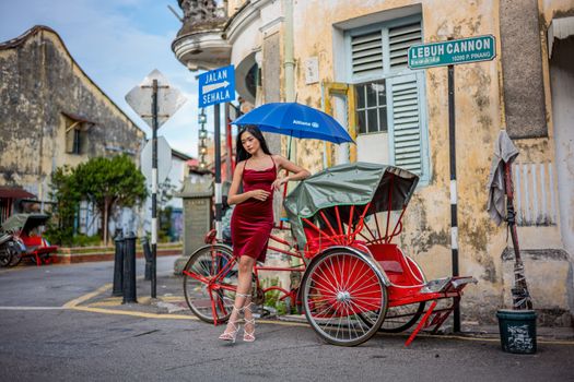 An elegant model in a red dress poses with a rickshaw on a vibrant street in George Town, Penang, Malaysia.