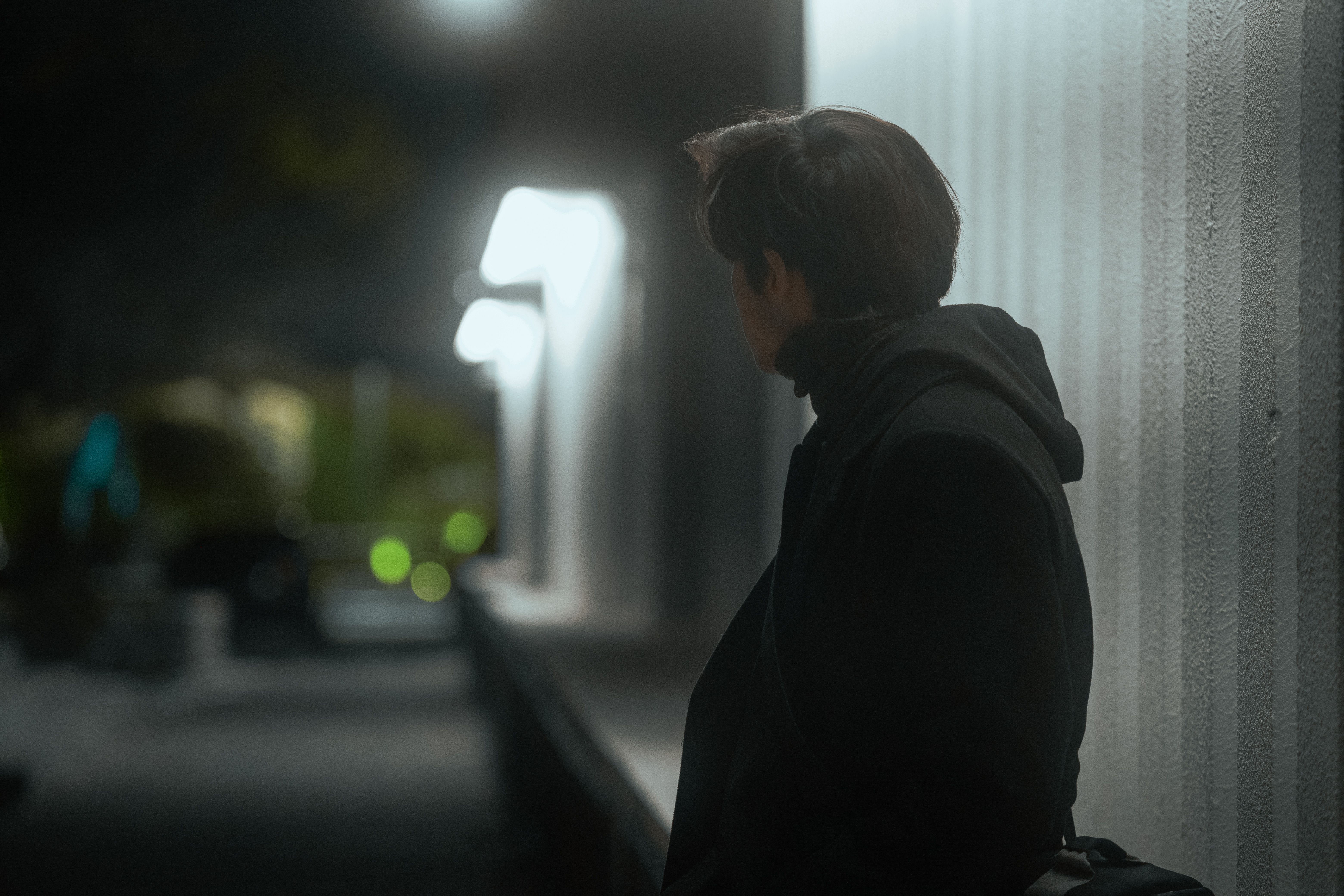 A person in a dark coat stands against a lit urban wall at night, creating a moody atmosphere.