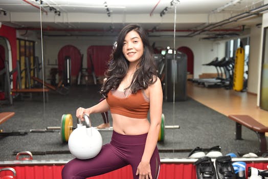 A smiling young woman in gym clothing holds a kettlebell indoors, embodying fitness and strength.