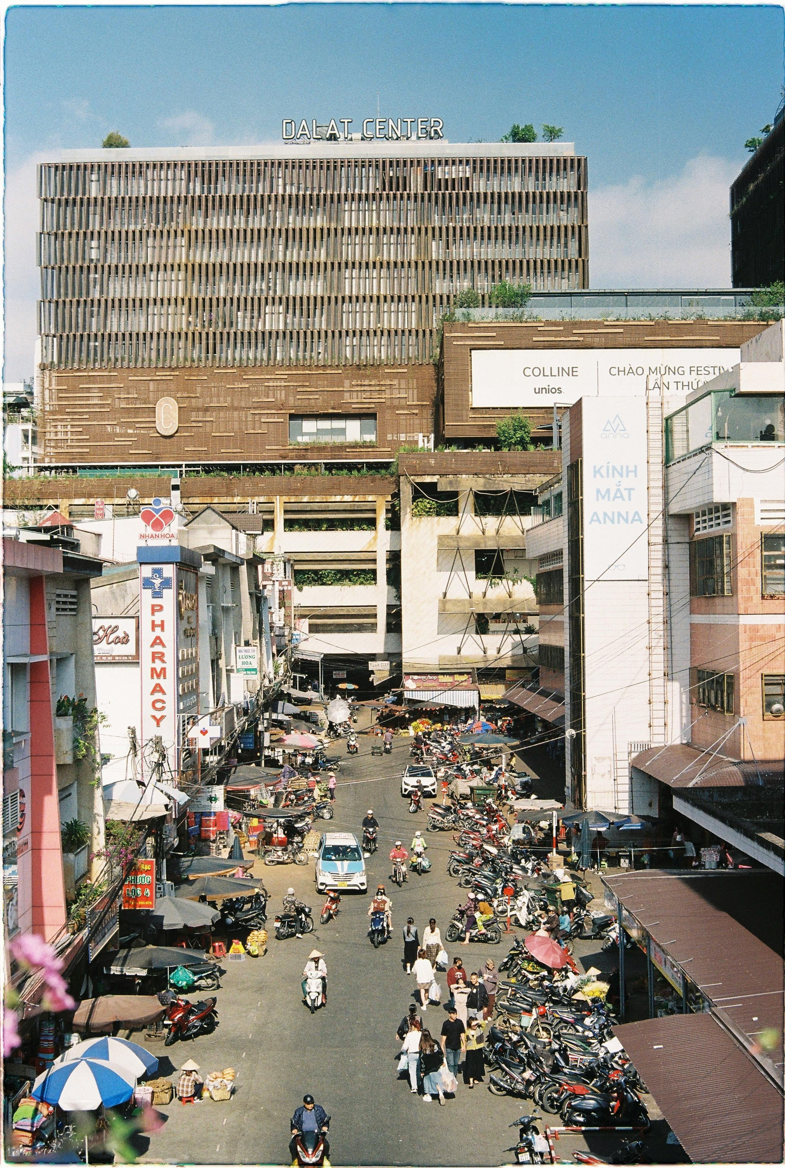 Vibrant marketplace in Dalat with people, motorbikes, and shops bustling under a clear sky.