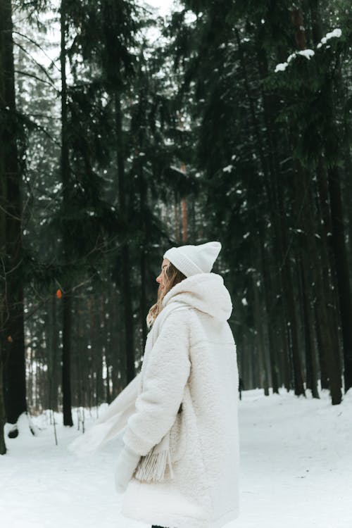 Blonde woman wears white fur coat and knitted hat posing with fir tree in winter forest. Winter fashion