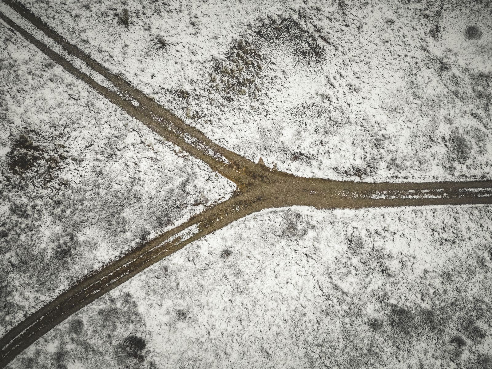 Aerial view straight down over a snow covered field where two dark muddy tracks come down from the top and merge into a single track heading right, with vehicle marks pressed into the white surface