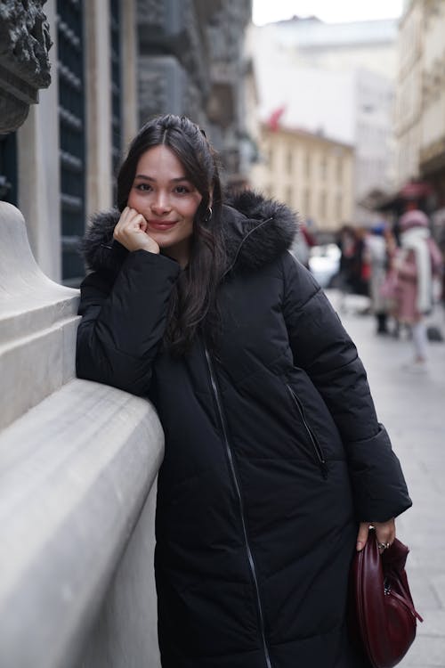 Young Woman in a Black Coat Standing by a Wall on a Sidewalk 