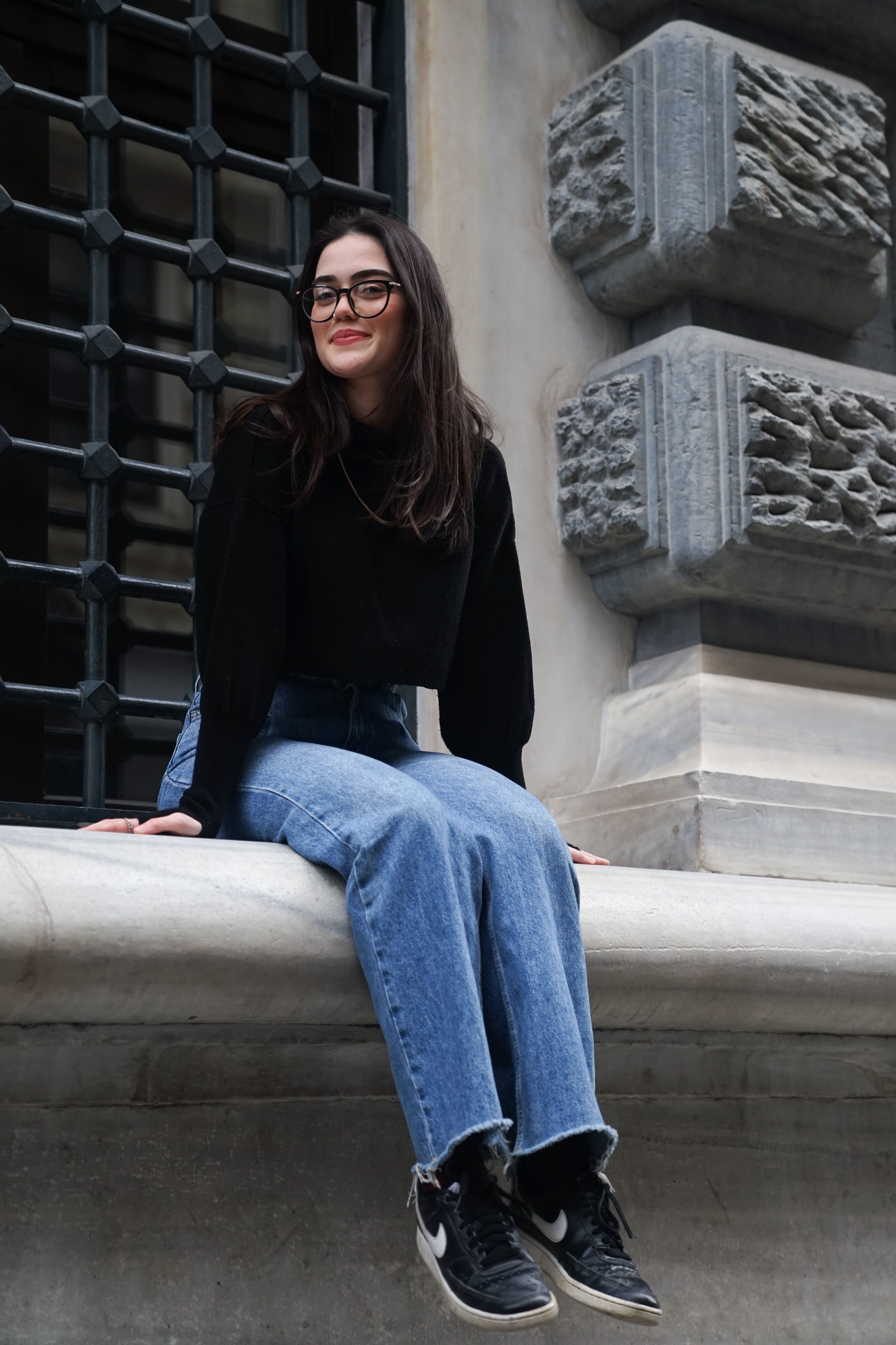 Fashionable woman in black sweater and jeans sitting outdoors on a windowsill with a smile.