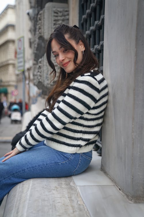 Young Woman in Jeans and Striped Sweater Sitting on a Wall in City 