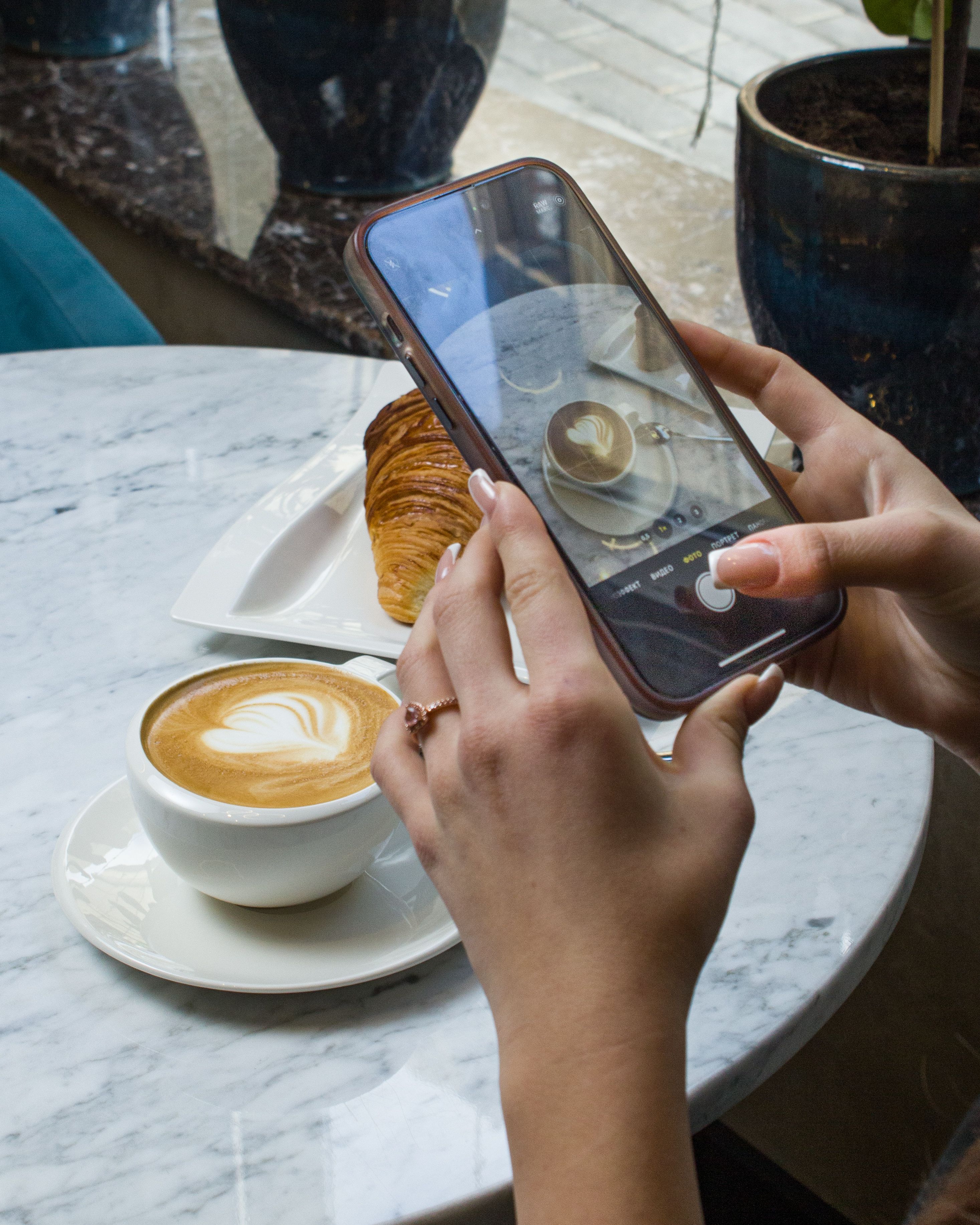 Close-up of a woman photographing latte art and a croissant in a cozy cafe with natural lighting.