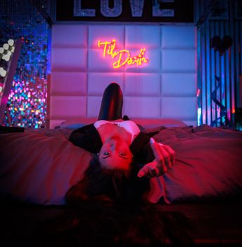 A woman in black outfit posing on a bed under vibrant neon lights, creating a moody and stylish ambiance.