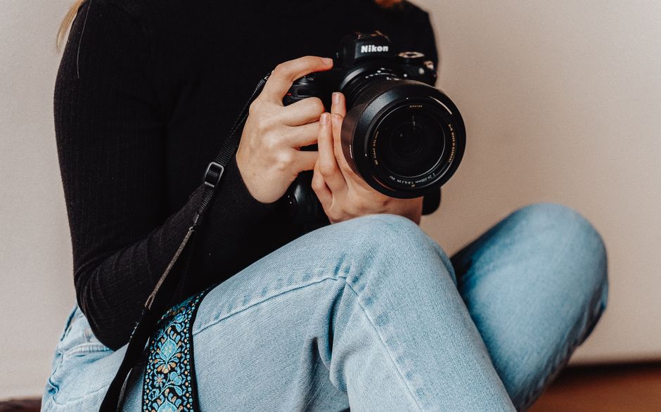 A woman holding a DSLR camera, seated casually indoors in blue jeans. A woman holding a DSLR camera, seated casually indoors in blue jeans.