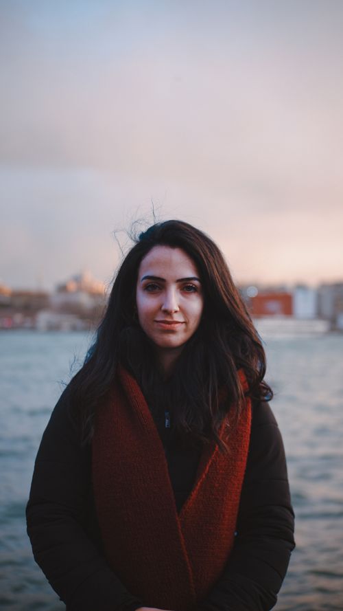 Free A stylish woman stands by the water during dusk, showcasing fall fashion with a scarf. Stock Photo