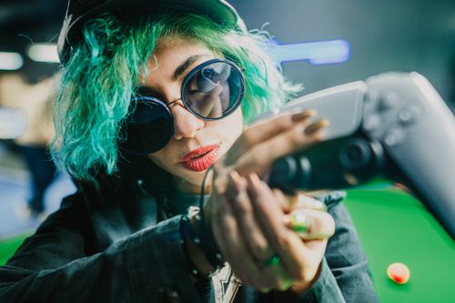 Free Edgy woman with green hair and sunglasses playing VR in a vibrant pub setting. Stock Photo