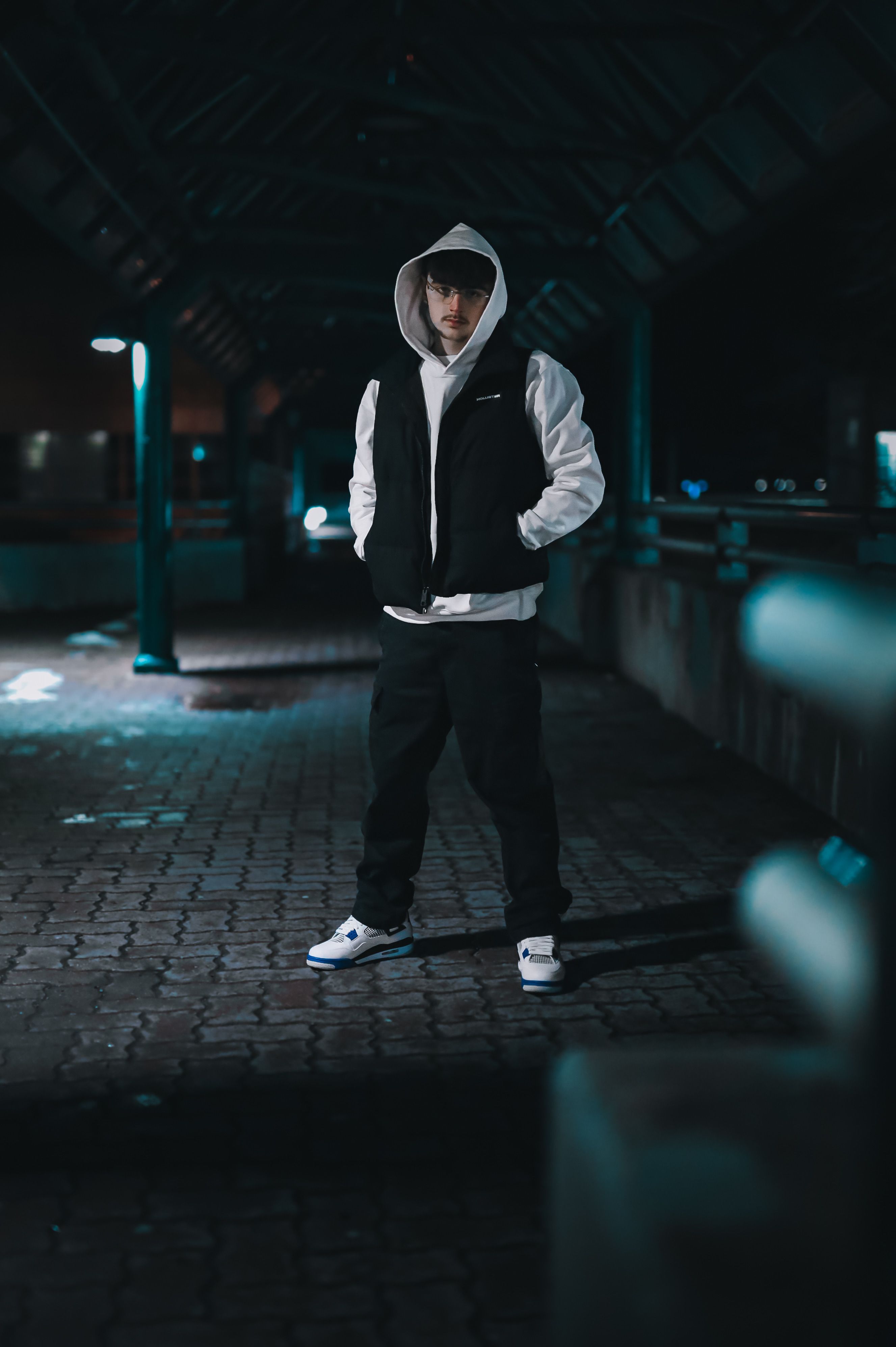 Stylish man in hoodie and sneakers stands confidently in dimly lit urban setting.