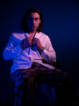 A musician in a white shirt poses in moody blue lighting with checkered pants.