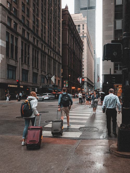 Free People crossing a bustling city street with luggage, surrounded by tall buildings and city life. Stock Photo