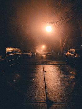 Moody view of a foggy city street at night with parked cars and streetlights.
