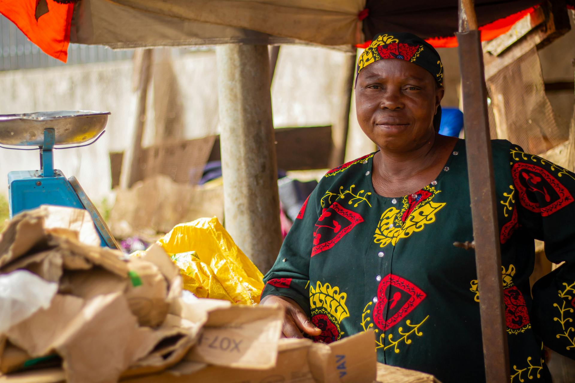 A smiling African woman vendor at her outdoor market stall surrounded by items and a weighing scale.