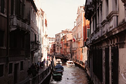 Explore a picturesque Venetian canal with boats and pedestrians strolling under the warm sun.