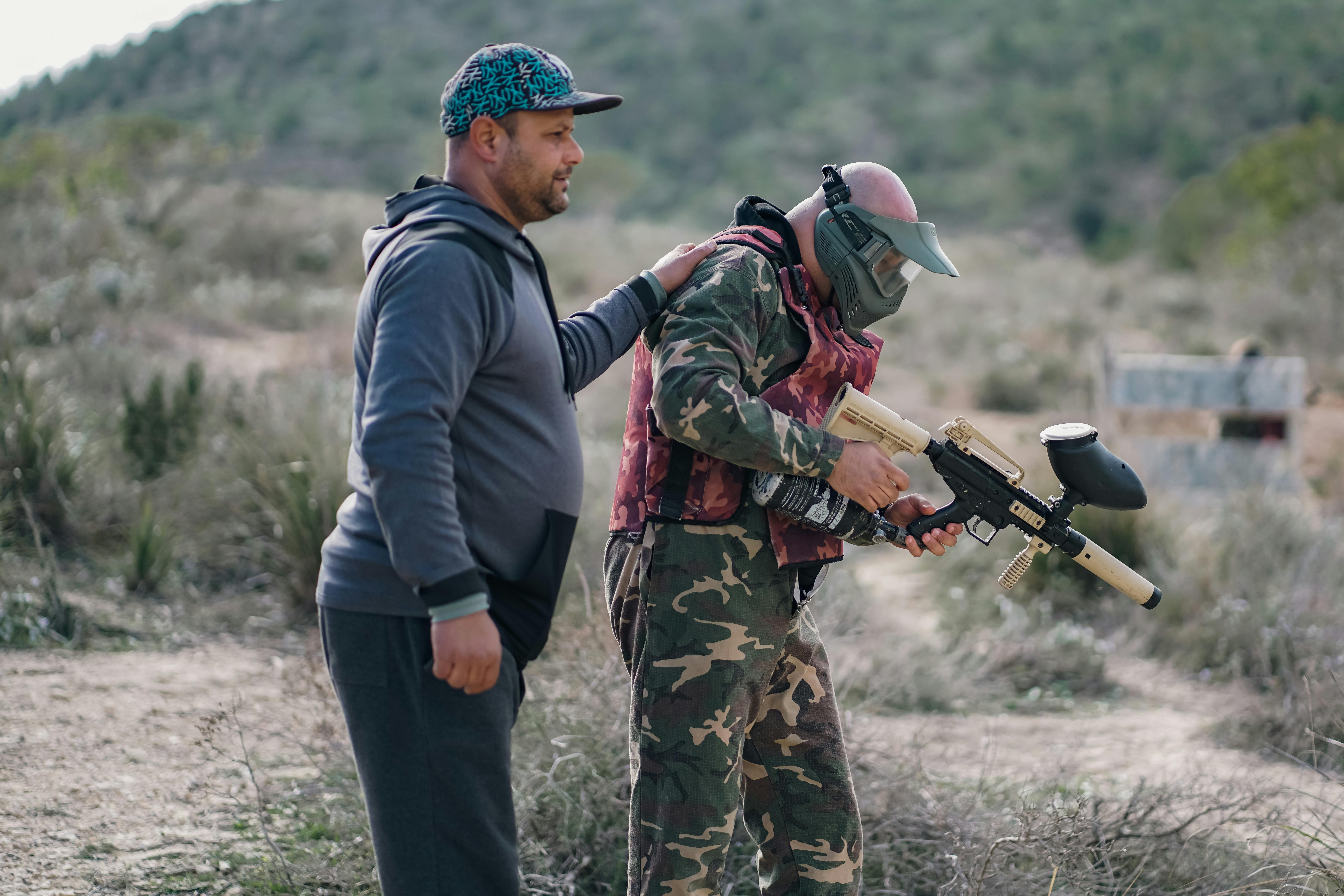 Two men playing paintball outdoors, one wearing camouflage and a mask, in a rural setting. Prescription Goggle Inserts - Two men playing paintball outdoors, one wearing camouflage and a mask, in a rural setting.