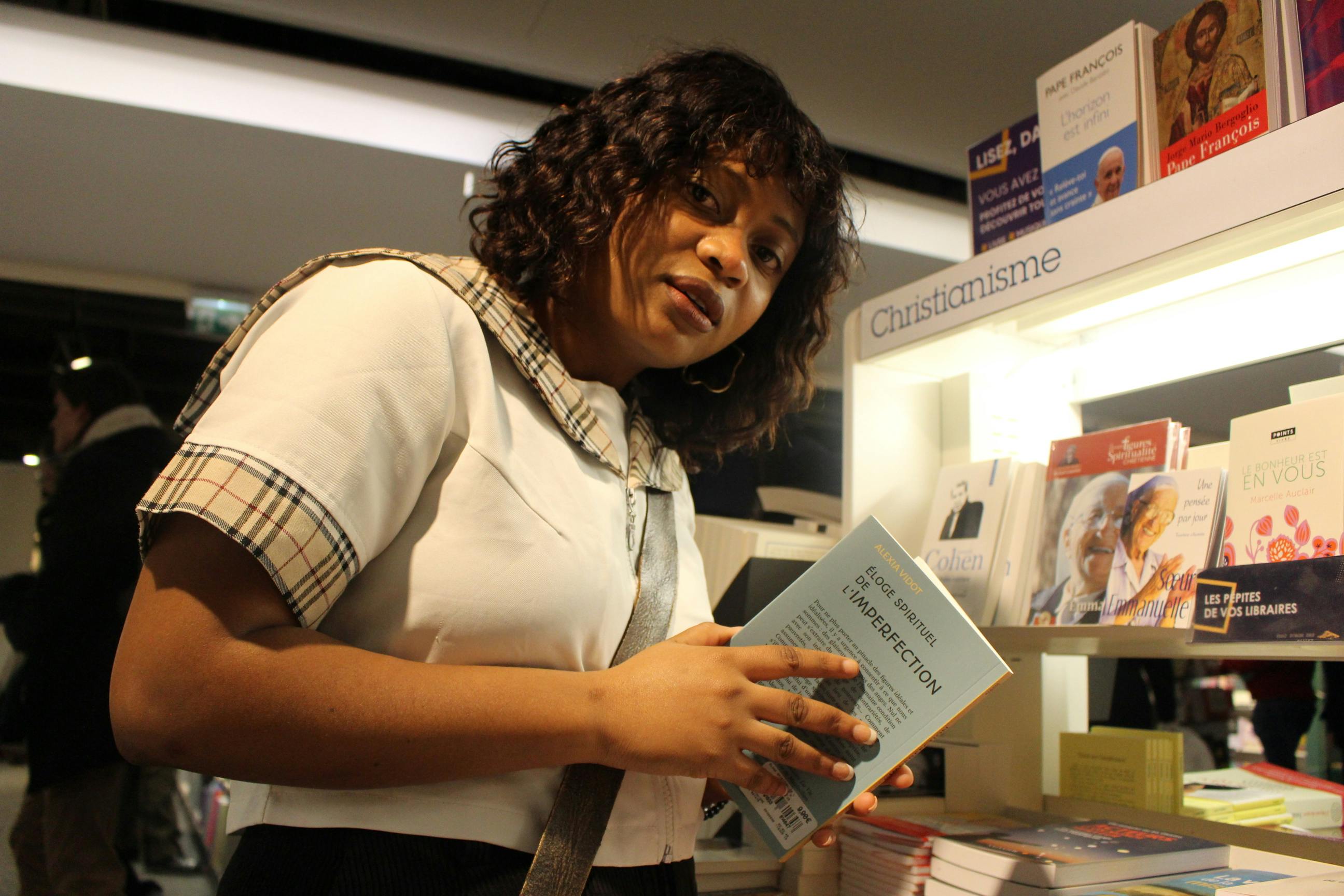 A woman is looking at a book in a section labeled 'Christianisme' within a bookstore. She appears engaged and thoughtful.