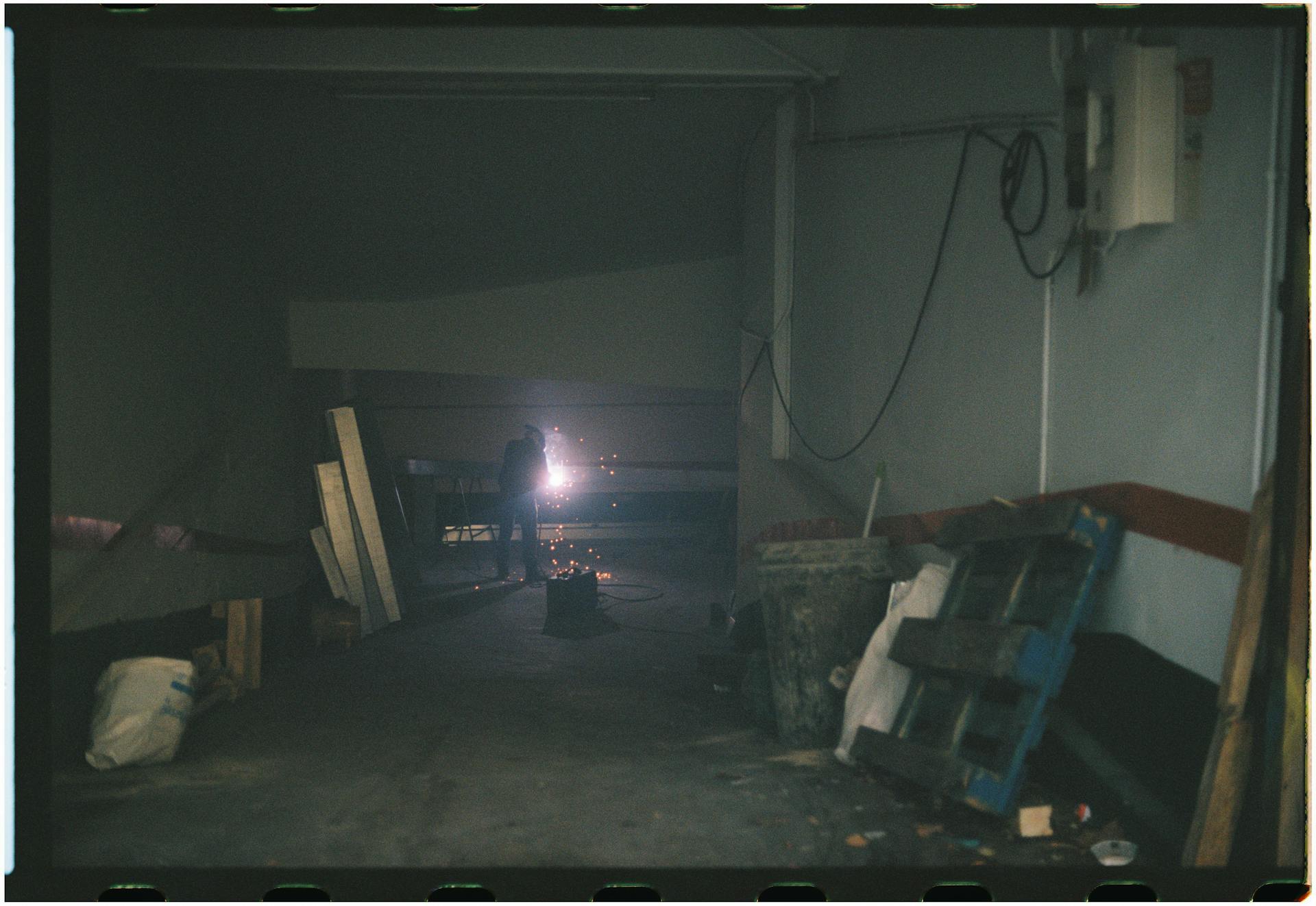 A single worker welding in a dimly lit garage filled with wooden pallets and debris.