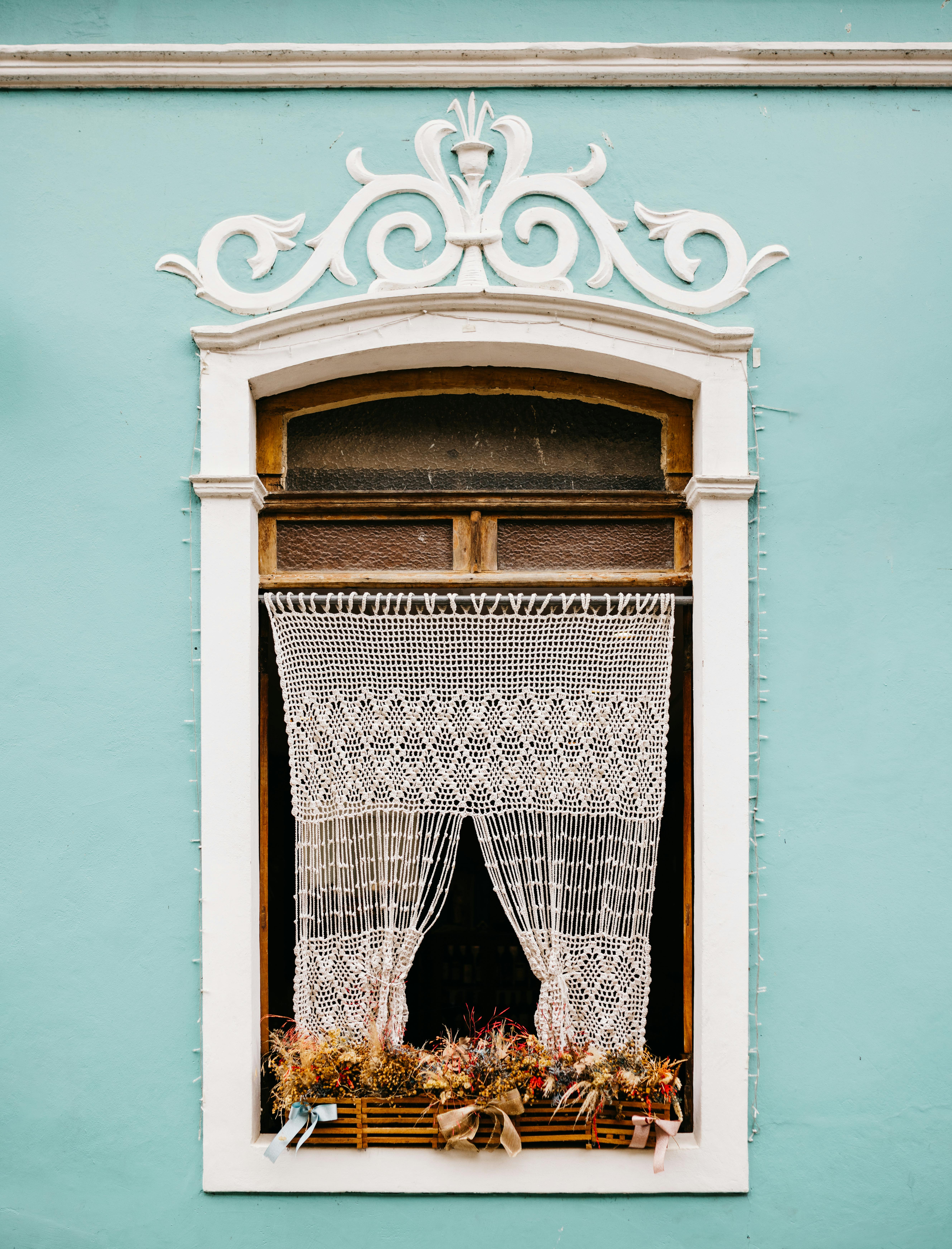 Charming teal wall with ornate window and lace curtains, showcasing vintage architectural style.