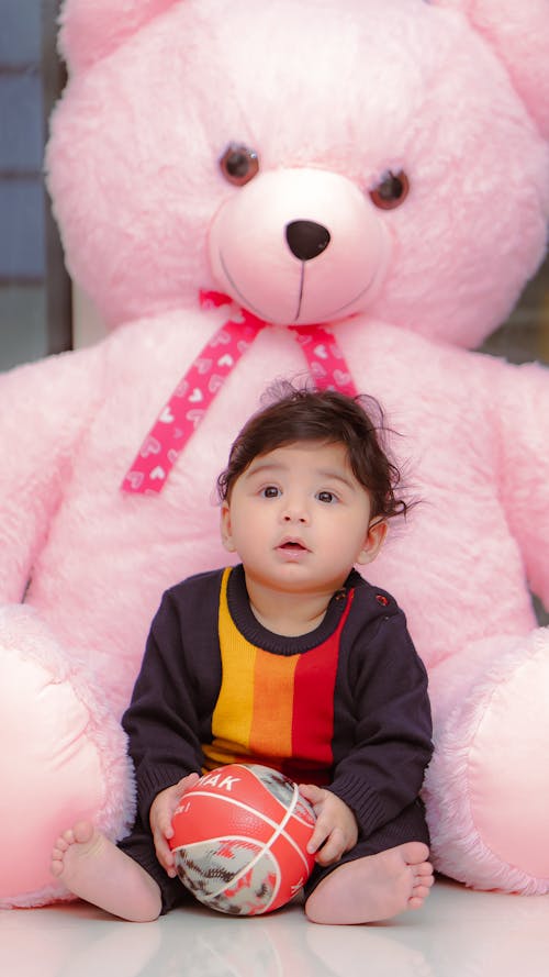 A baby sitting on the floor next to a large pink teddy bear