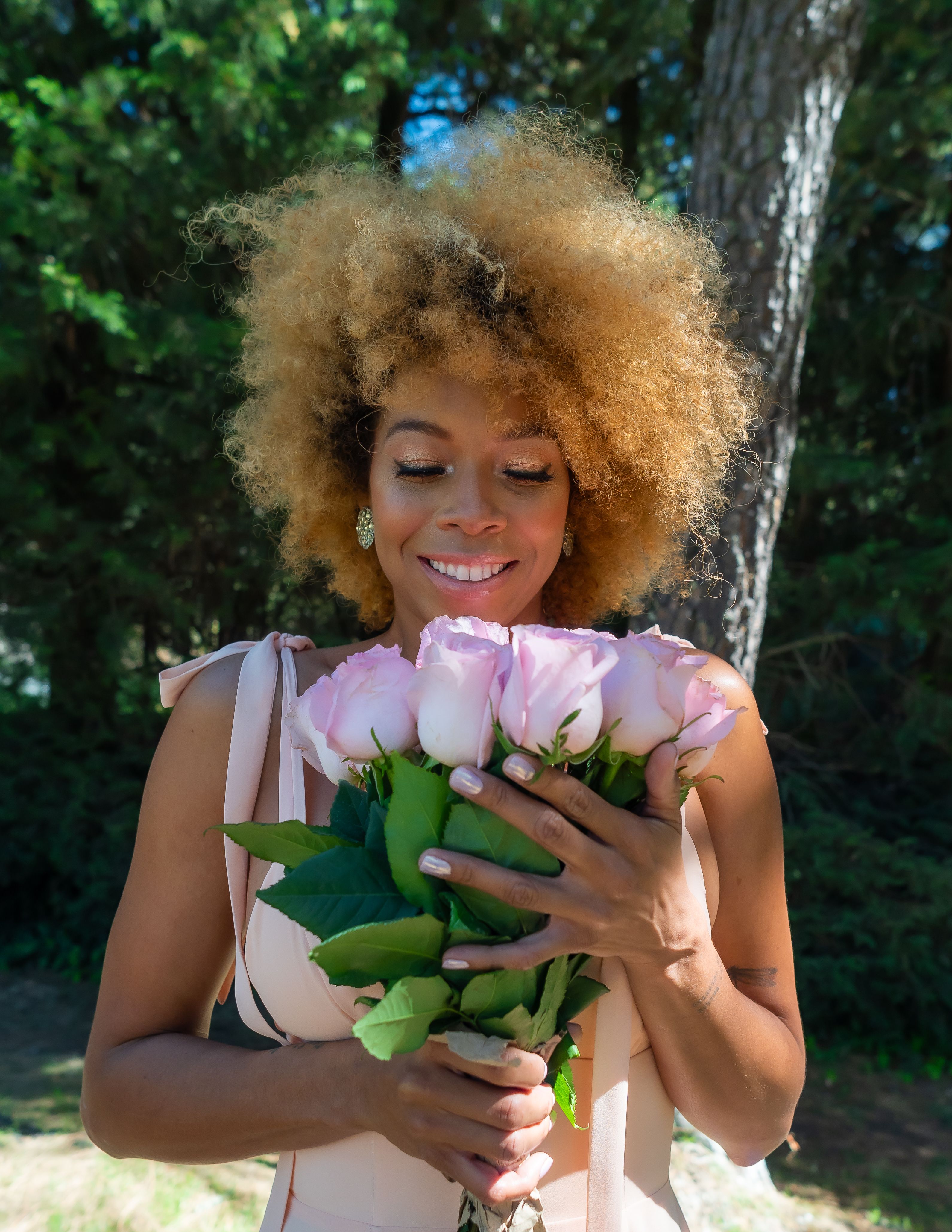 A cheerful woman with natural afro hair holds pink roses outdoors in a sunny garden, CDMX, Mexico.