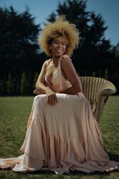 A stylish woman with afro hair sitting outdoors, smiling in a flowing peach dress, exuding elegance.