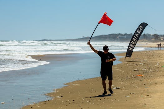A man walks along the beach holding a red flag, signifying danger or caution near the ocean shore.