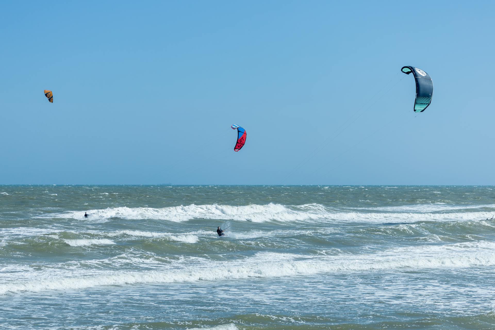 Three kitesurfers riding the waves in a clear blue sky, capturing a fun and adventurous moment.