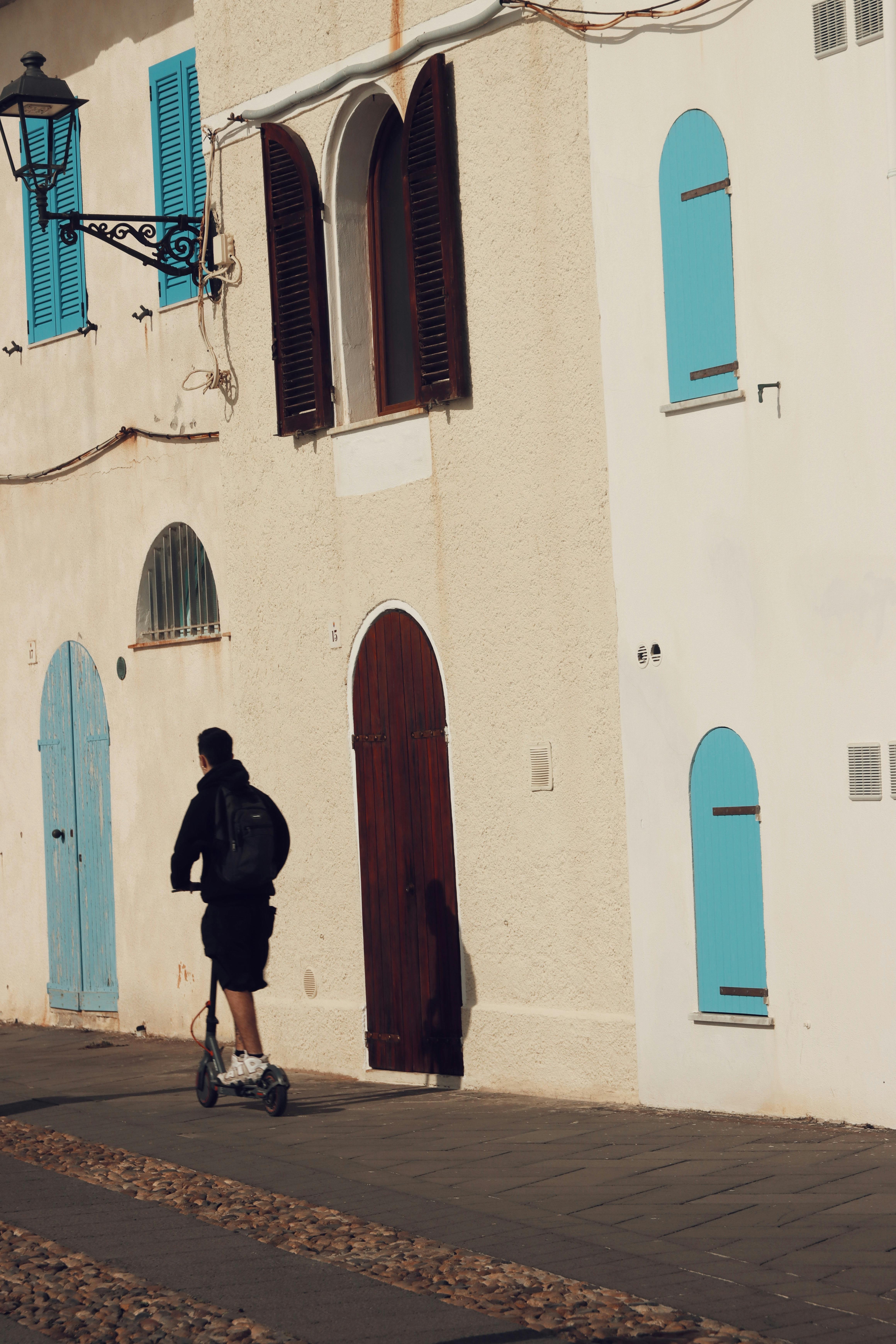 A person rides a scooter past colorful shutters in Alghero, Italy, highlighting vibrant street life.