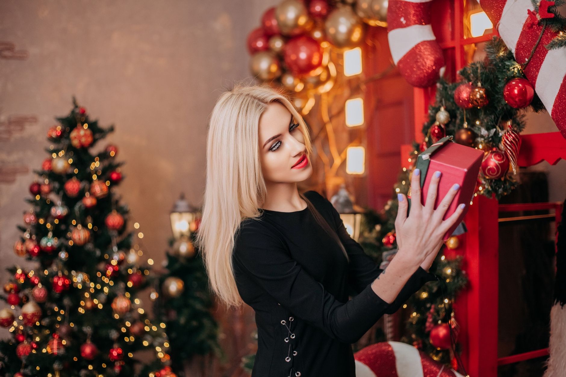 Blonde woman in black dress holding a Christmas present with festive decor.